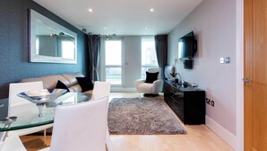 A modern living room with a glass coffee table, a sofa, a TV, and a large mirror on the wall.