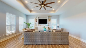 A living room with a ceiling fan, a large flat-screen TV, wicker furniture, and a potted plant.