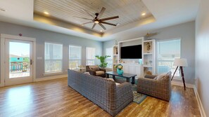 A spacious living room with a ceiling fan, a flat-screen TV, wicker furniture, and a view through large windows. - Adicora Beach House: 3 BR, 2 BA House in Port Bolivar, Sleeps 14 (Bolivar Peninsula)