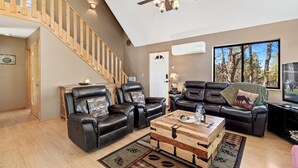 A spacious living room with a wooden staircase, leather sofas, a wooden coffee table, and a large window with a view of trees. - High Country Escape | Forest Border • Fenced Yard (Heber)