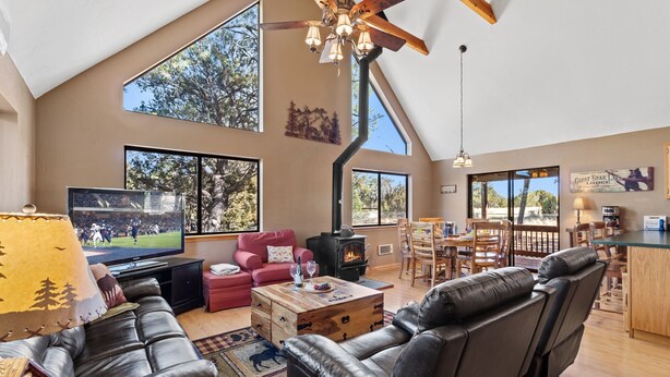 A spacious living room with a large TV, leather sofas, a fireplace, and a dining area with wooden furniture. - High Country Escape | Forest Border • Fenced Yard (Heber)