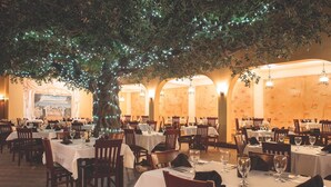 Um restaurante com mesas redondas, toalhas brancas e cadeiras. Há uma grande árvore com luzes no centro.