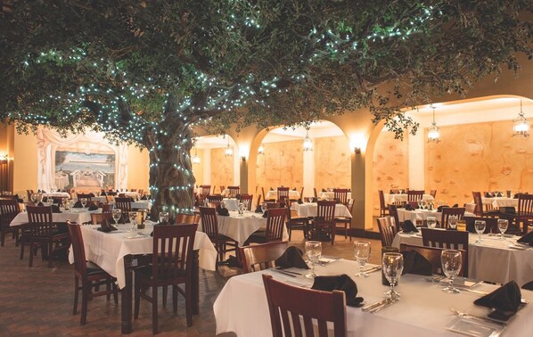 Un restaurante con mesas redondas, manteles blancos y sillas. Hay un gran árbol con luces en el centro.