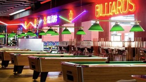 Uma sala de bilhar com várias mesas, letreiros de neon e luminárias pendentes verdes.