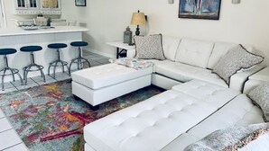 Uma sala de estar moderna com um sofá seccional branco, um tapete colorido e uma área de cozinha com armários brancos e banquetas.