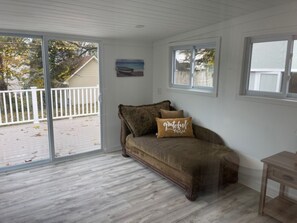 A room with a sliding glass door leading to a deck, a brown sofa with a 'grateful' pillow, and a small wooden table. - Haven Breeze - Dazzling home near Lake Ontario, beach & relaxing walking trails (Rochester)