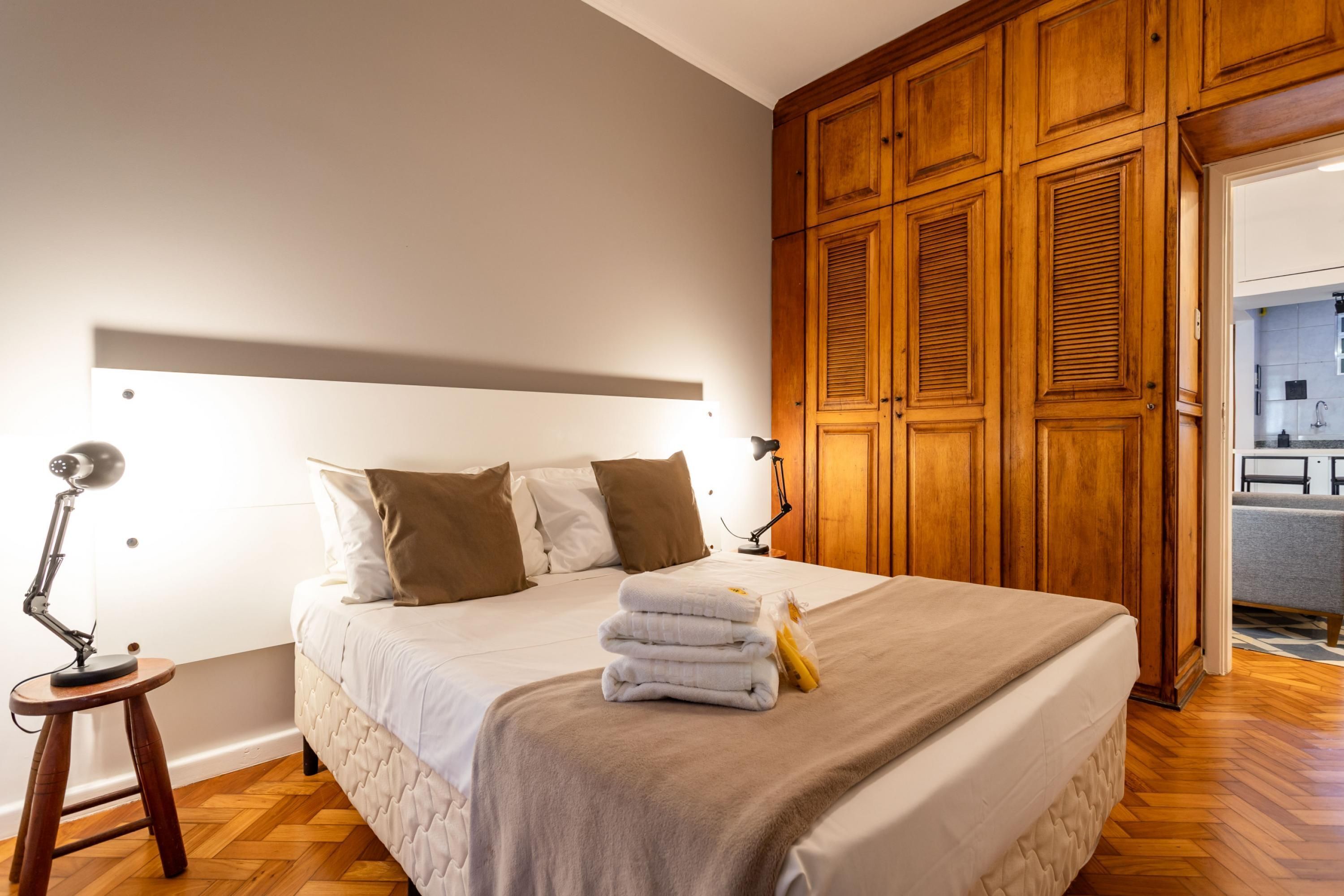 Een net opgemaakt bed met hoofdbord, een bedlampje en een stapje opgevouwen handdoeken.