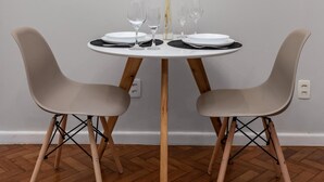 Et spiseområde med et rundt bord dækket til to, omkranset af to stole, en vase med tørrede planter og tre billeder på væggen.