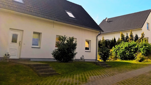Een twee verdiepingen tellend huis met een schuin dak, een voordeur en een klein raam. Een verhard pad leidt naar de voordeur.