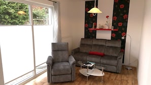 Een woonkamer met een grijze bank, een grijze fauteuil, een kleine witte salontafel en een vloerlamp. Er is een raam met uitzicht op groen.