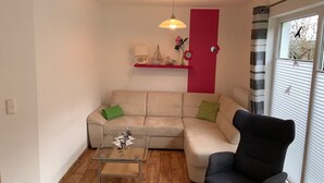 Ein Wohnzimmer mit einem Glastisch, einem beige-farbenen Sofa mit grünen Kissen, einem schwarzen Sessel, einer roten Akzentwand und einer hängenden Pendelleuchte.