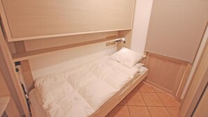 Ein kleines Zimmer mit einem Einzelbett, einem Holzkopfstück, einem Fenster mit Jalousien und einem Fliesenboden.