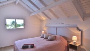 A bedroom with a large bed, a nightstand, and a window with a view of palm trees. - Éden love! (Collectivité de Saint-Martin)