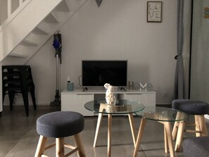 Un saló modern amb televisió de pantalla plana, una taula de cafè de vidre i dos tamborets amb coixins.