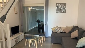 A modern living room with a grey sofa, a glass coffee table, and a TV on a white stand. There is a staircase on the left and a door leading to another room. - Éden love! (Collectivité de Saint-Martin)