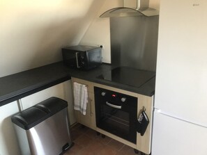 Une petite cuisine équipée avec un plan de travail noir, un four en acier inoxydable, une petite poubelle et un réfrigérateur blanc.