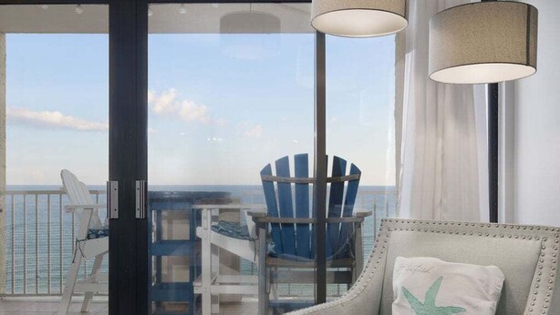 A balcony with a view of the ocean, featuring two blue Adirondack chairs and a white armchair with a starfish pillow. - Tropical Winds 603 2 Bedroom Condo by RedAwning (Gulf Shores)