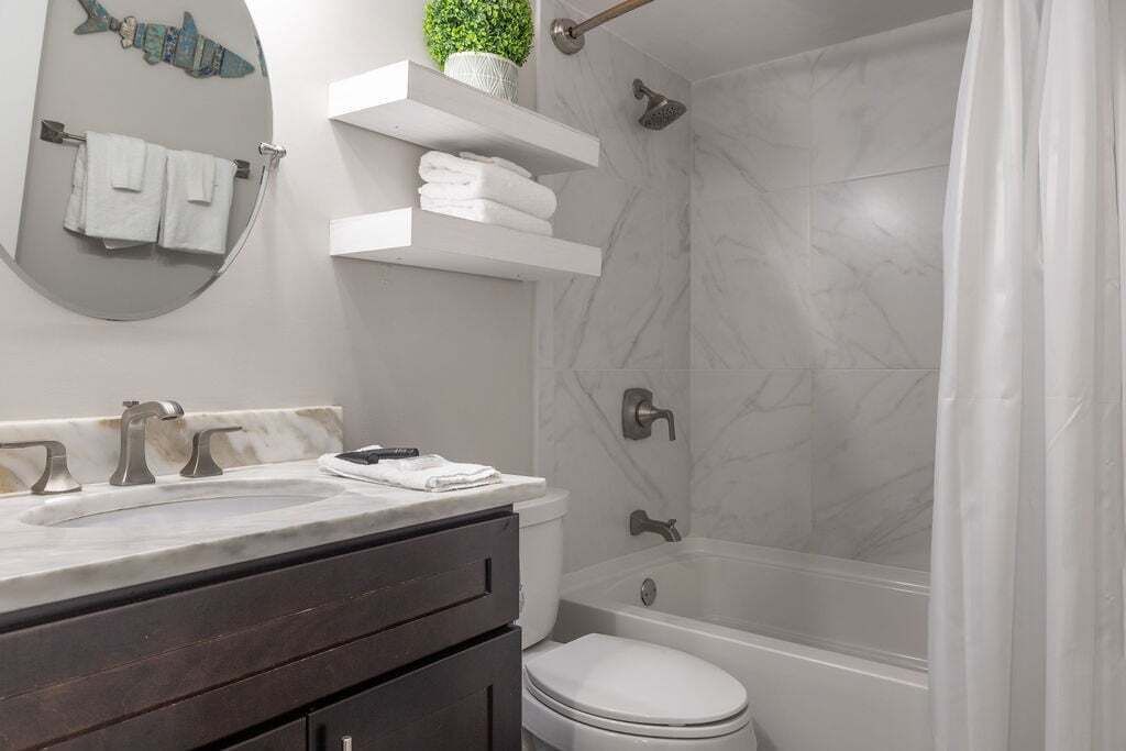 Une salle de bain moderne avec un lavabo en marbre, des toilettes, une baignoire et une douche. On y trouve des serviettes blanches et une plante en pot sur les étagères.