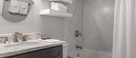 Une salle de bain moderne avec un lavabo en marbre, des toilettes, une baignoire et une douche. On y trouve des serviettes blanches et une plante en pot sur les étagères.