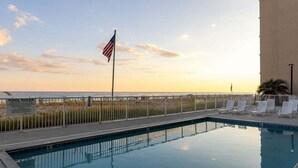 A pool with a view of the ocean at sunset, a flag, and lounge chairs. - Tropical Winds 603 2 Bedroom Condo by RedAwning (Gulf Shores)
