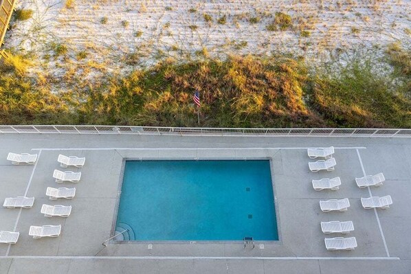 Une piscine rectangulaire entourée de chaises longues blanches, un drapeau et un mur de béton agrémenté de quelques plantes.