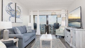 A modern living room with a grey sofa, white coffee table, and a view of a balcony with chairs. - Tropical Winds 603 2 Bedroom Condo by RedAwning (Gulf Shores)