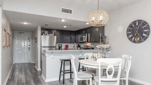 A modern kitchen with dark cabinets, a white island, and a dining area with white chairs. - Tropical Winds 603 2 Bedroom Condo by RedAwning (Gulf Shores)
