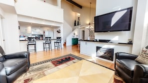 Un amplio salón con un televisor grande, sofás de cuero, un tapete estampado y una cocina al fondo.