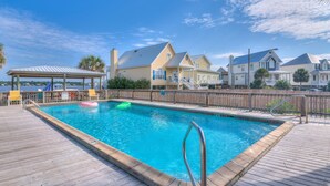 A swimming pool with a wooden deck, a covered area with chairs, and houses in the background. - Lagoon Landing 305 1 Bedroom Condo by RedAwning (Gulf Shores)
