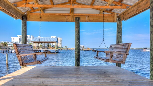 A wooden deck with two swings over water, a covered area, and buildings in the background. - Lagoon Landing 305 1 Bedroom Condo by RedAwning (Gulf Shores)