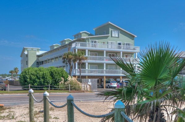 A multi-story hotel with balconies, a parking area, and a sandy foreground with palm trees. - Lagoon Landing 305 1 Bedroom Condo by RedAwning (Gulf Shores)