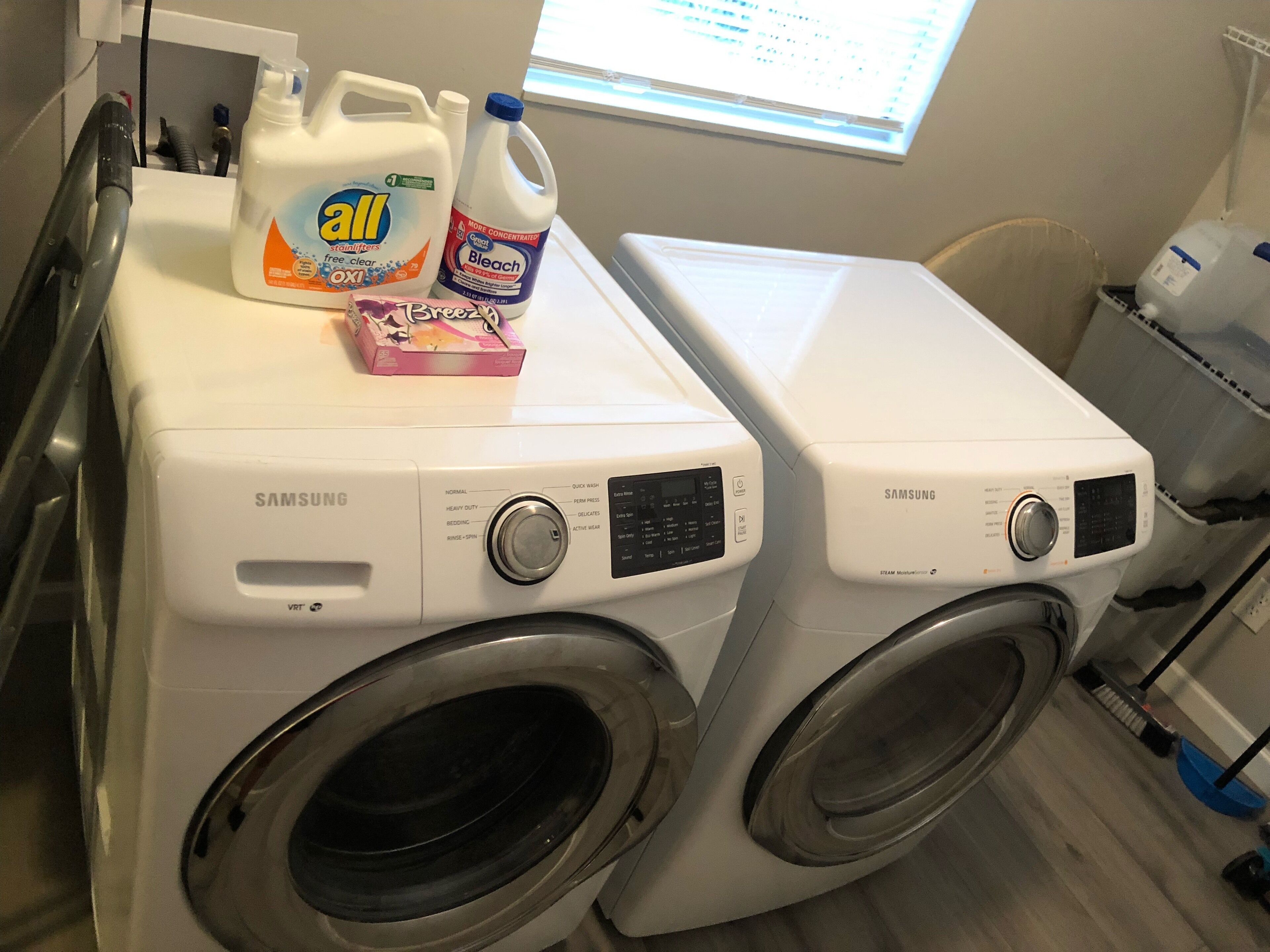 A laundry area with two Samsung washing machines, a bottle of All detergent, and a bottle of bleach.