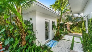 Um edifício branco com porta de vidro, rodeado por vegetação exuberante e um caminho de azulejos.