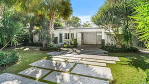 Uma casa moderna com um caminho de pedras que leva à entrada, rodeada por vegetação exuberante e palmeiras.