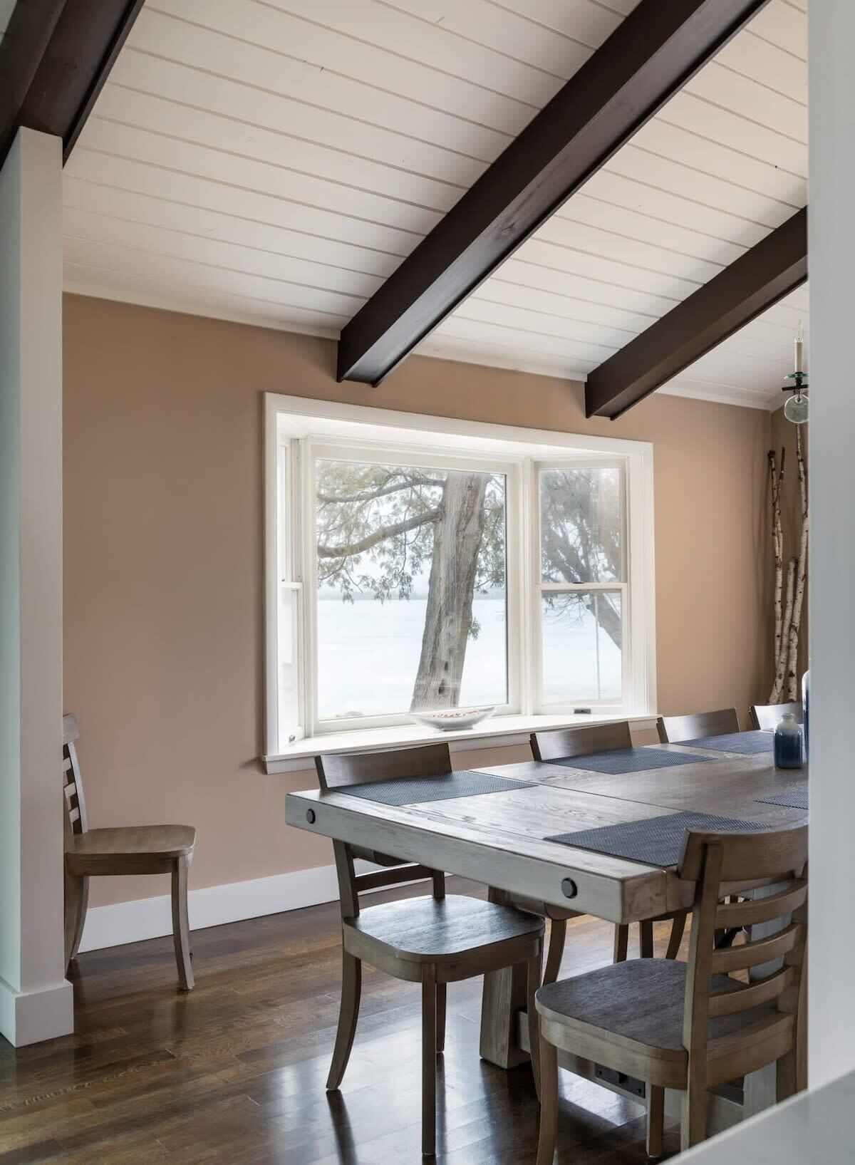 A dining area with a wooden table and chairs, a window with a view of trees, and a ceiling with exposed beams.