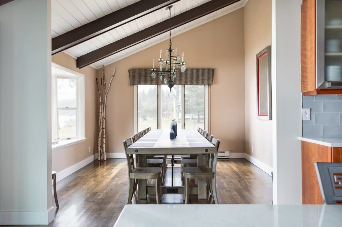 A dining area with a long table, chairs, a chandelier, and a view through large windows.