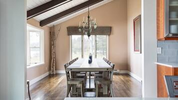 A dining area with a long table, chairs, a chandelier, and a view through large windows.