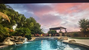 Una piscina con una cascada, rodeada de rocas y vegetación. Se observan sillas de descanso y un pabellón al fondo.
