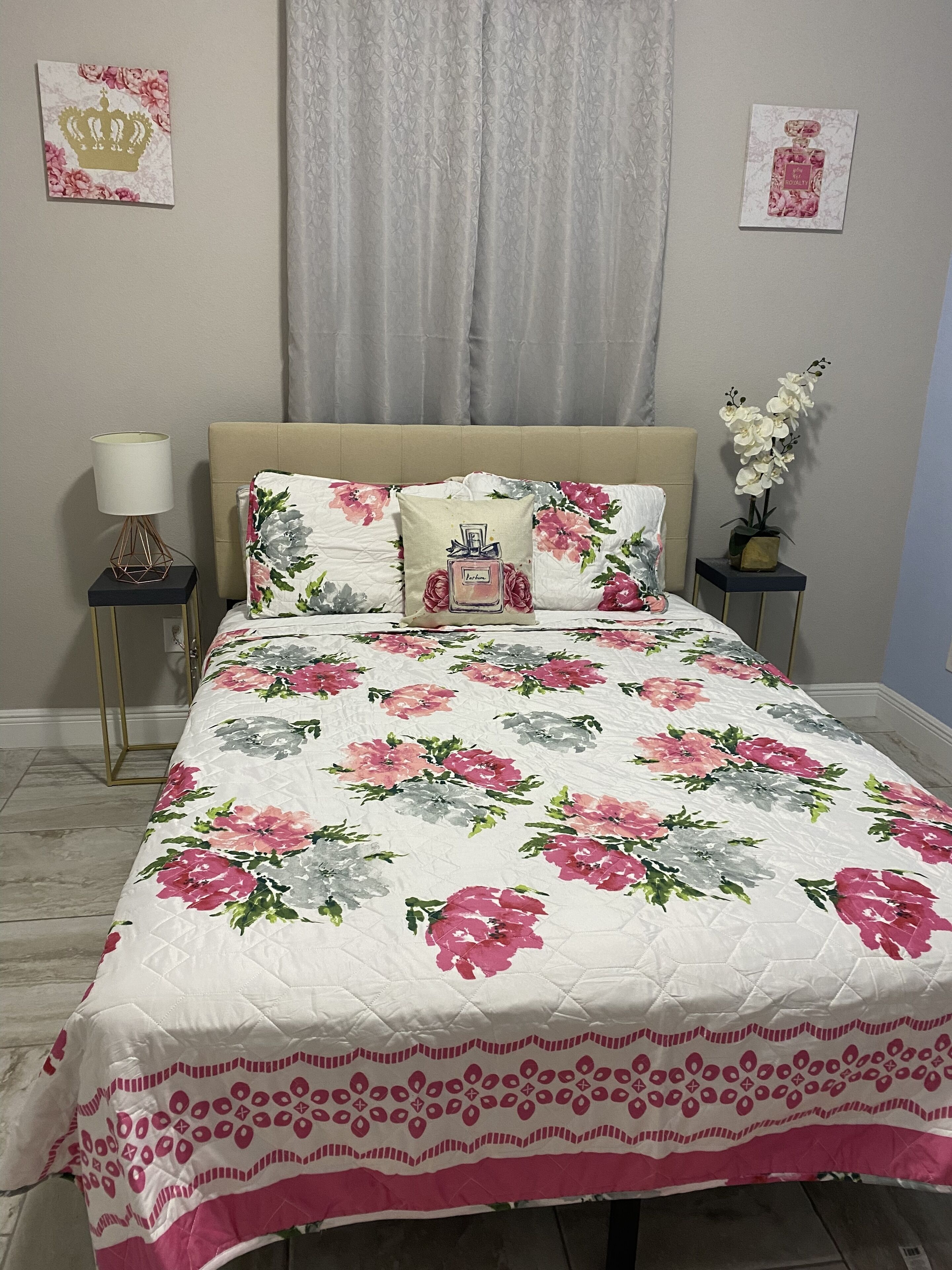 Un dormitorio con una colcha estampada de flores, un cabecero color beige, una mesita de noche con lámpara y una pequeña planta en maceta.