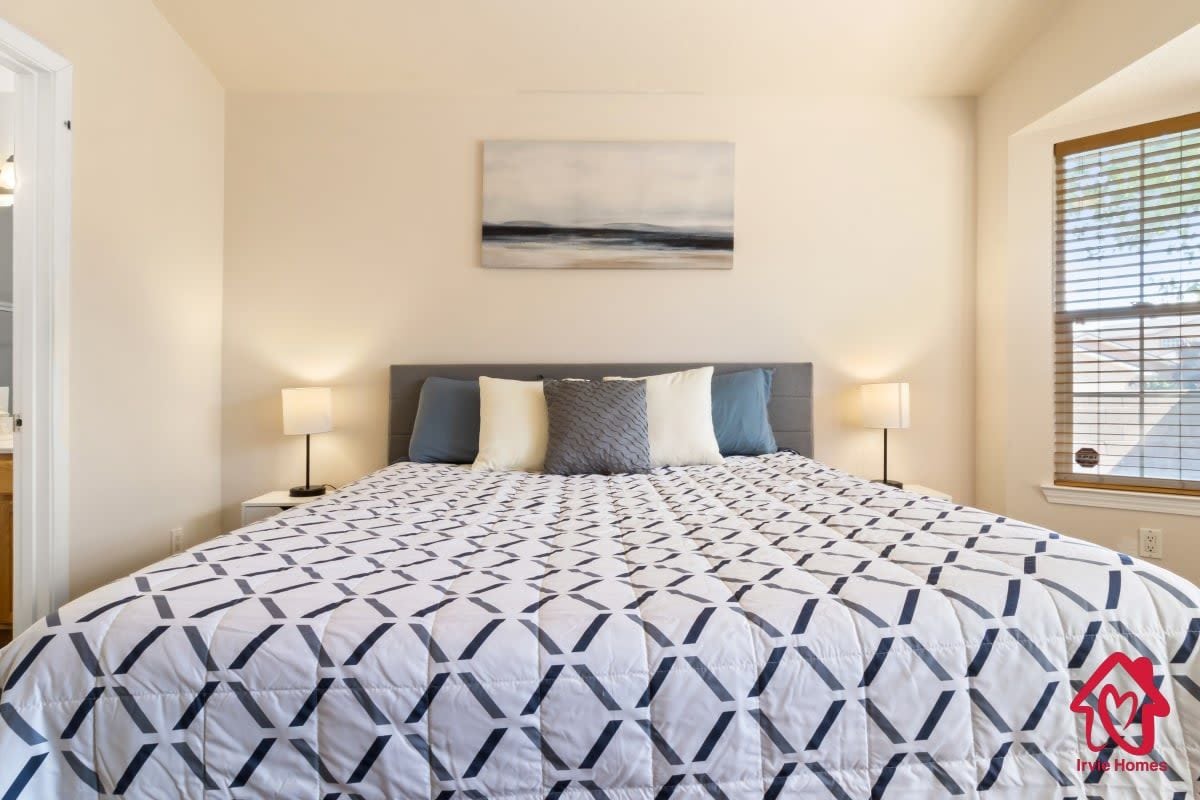 A bedroom with a bed, two bedside lamps, a wall-mounted painting, and a window with blinds.