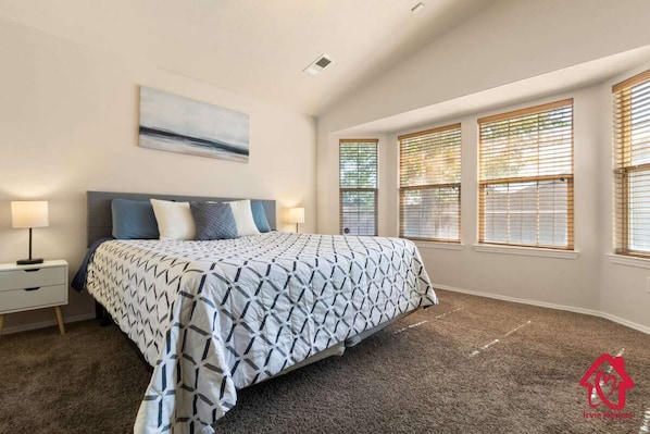 A bedroom with a large bed, a nightstand, a wall-mounted lamp, and three windows with blinds.