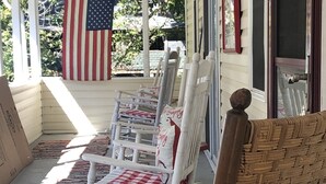 Eine Veranda mit weißen Schaukelstühlen und einem Korbstuhl, einer rot-weiß karierten Tischdecke und einer amerikanischen Flagge.