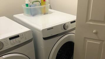 A laundry area with two white washing machines, one with a control panel and the other with a front-loading door. A plastic container with cleaning supplies is on top of the machine with the control panel.