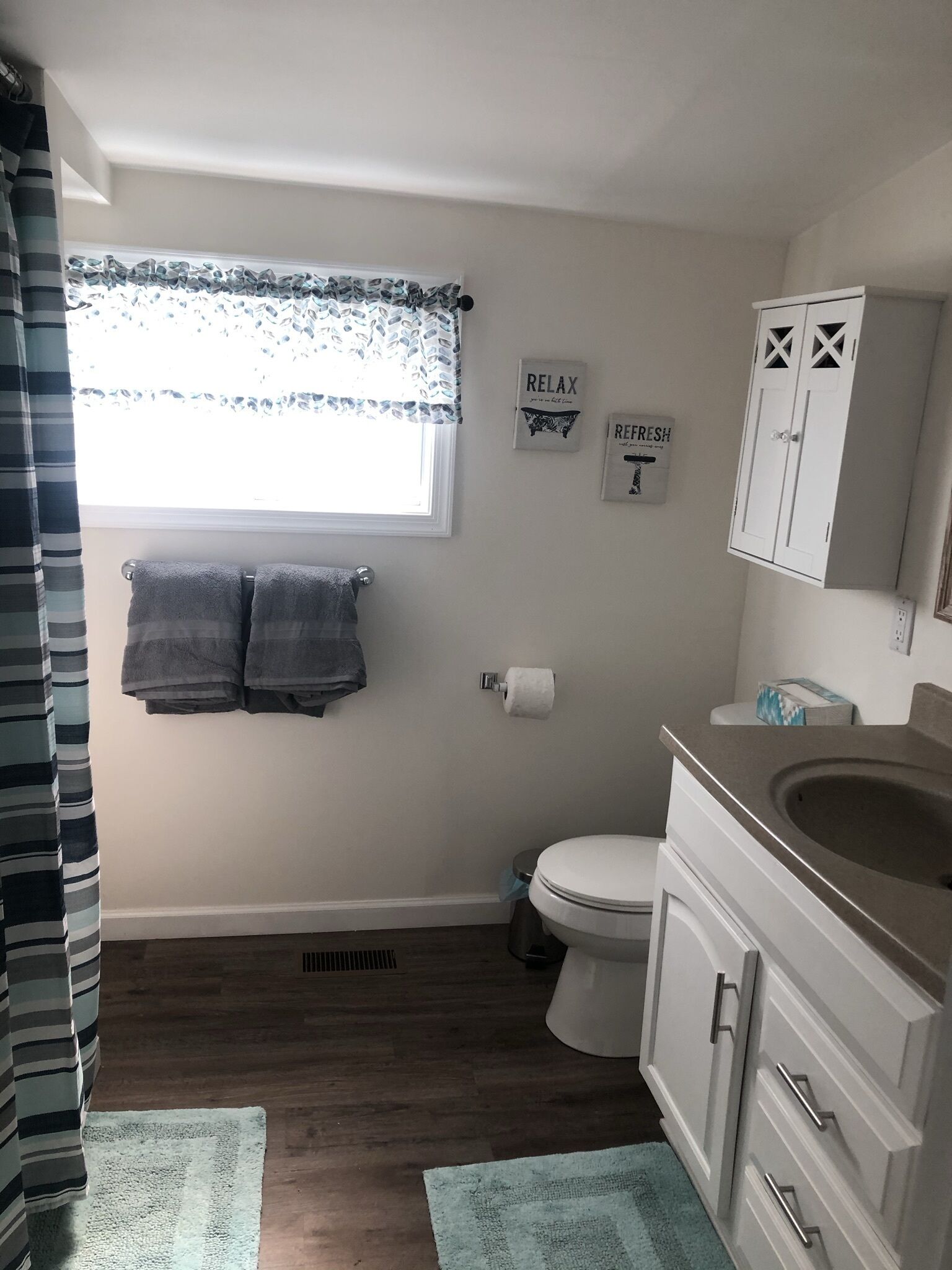 Une salle de bain comprenant des toilettes, un lavabo et un meuble de rangement, une fenêtre avec un rideau et deux serviettes accrochées au mur.