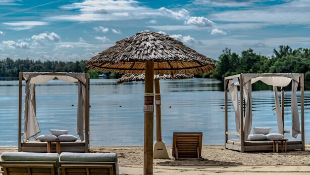 Ein Strand mit zwei Holzhütten, jede mit einem Reetdach und Kissen, mit Blick auf einen ruhigen See.