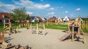 A playground with various wooden structures and a sandy area, surrounded by houses with different architectural styles.
