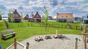 A playground with sand and wooden play structures, surrounded by a fence and houses in the background.