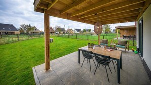 A covered patio area with a wooden table and chairs, a hanging light fixture, and a view of a green lawn and houses in the background.