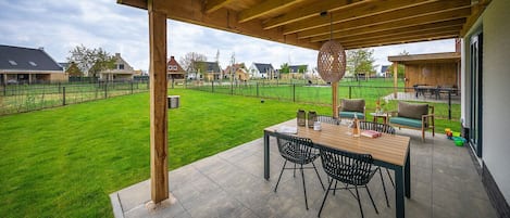 A covered patio area with a wooden table and chairs, a hanging light fixture, and a view of a green lawn and houses in the background.