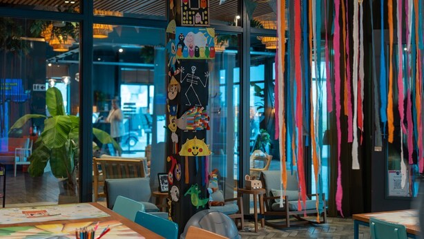 A brightly lit indoor space with large windows, a central column decorated with various items, and a seating area with tables and chairs.
